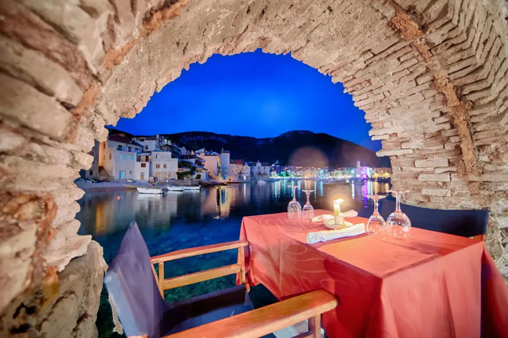 Romantic dining table with a red tablecloth and candles, set along a harbor framed by an ancient stone arch at dusk.
