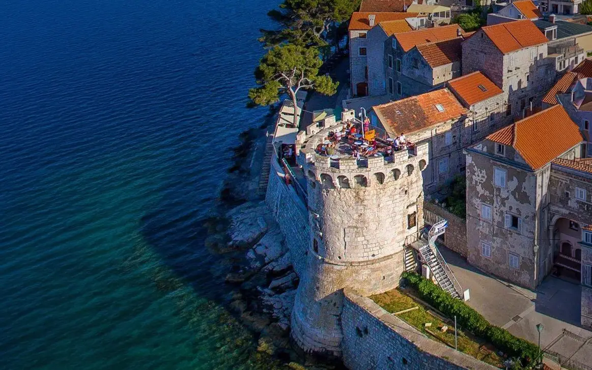 Aerial view of a stone fortress tower overlooking a turquoise sea, with orange-roofed houses along the coast and visitors on the terrace.
