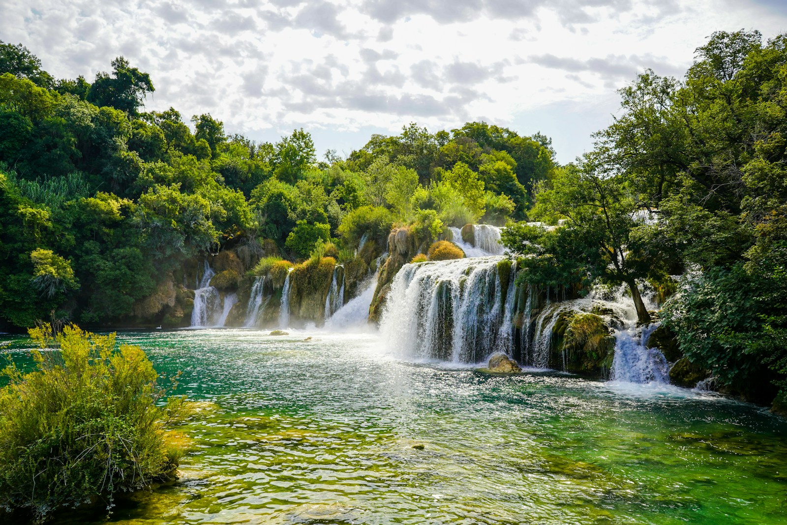 waterfalls near green trees under white clouds during daytime